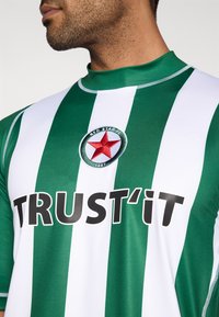 Zielono-biała koszulka w pionowe paski z wysokim kołnierzem, dużym czarnym napisem "TRUST'IT" oraz okrągłym czerwonym logo z gwiazdą.