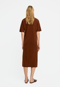 Brązowa sukienka do kolan z miękkiej dzianiny, z krótkimi rękawami i prostym krojem. Noszona w połączeniu z brązowymi butami wsuwanymi.