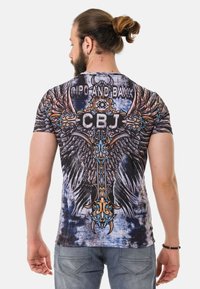 Grafisches T-Shirt mit einem großen Designe eines Adlerflügels mit verzierten Details und einem zentralen Kreuz. Der Stoff ist dehnbar und hat einen gemischten blau-grauen Hintergrund.