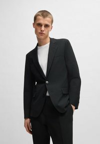 Blazer nero con texture, chiusura a un bottone, colletto a revers e due tasche frontali, indossato sopra una camicia bianca in un ambiente minimalista.