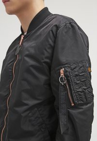 Czarna kurtka bomberka wykonana z gładkiego, błyszczącego materiału. Posiada zamek błyskawiczny z przodu, prążkowany kołnierz oraz kieszeń użytkową z zamkiem na rękawie.