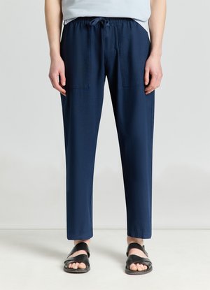 Persona che indossa pantaloni blu navy con coulisse e tasche applicate sul davanti e sandali neri con punta aperta, in piedi su uno sfondo semplice.