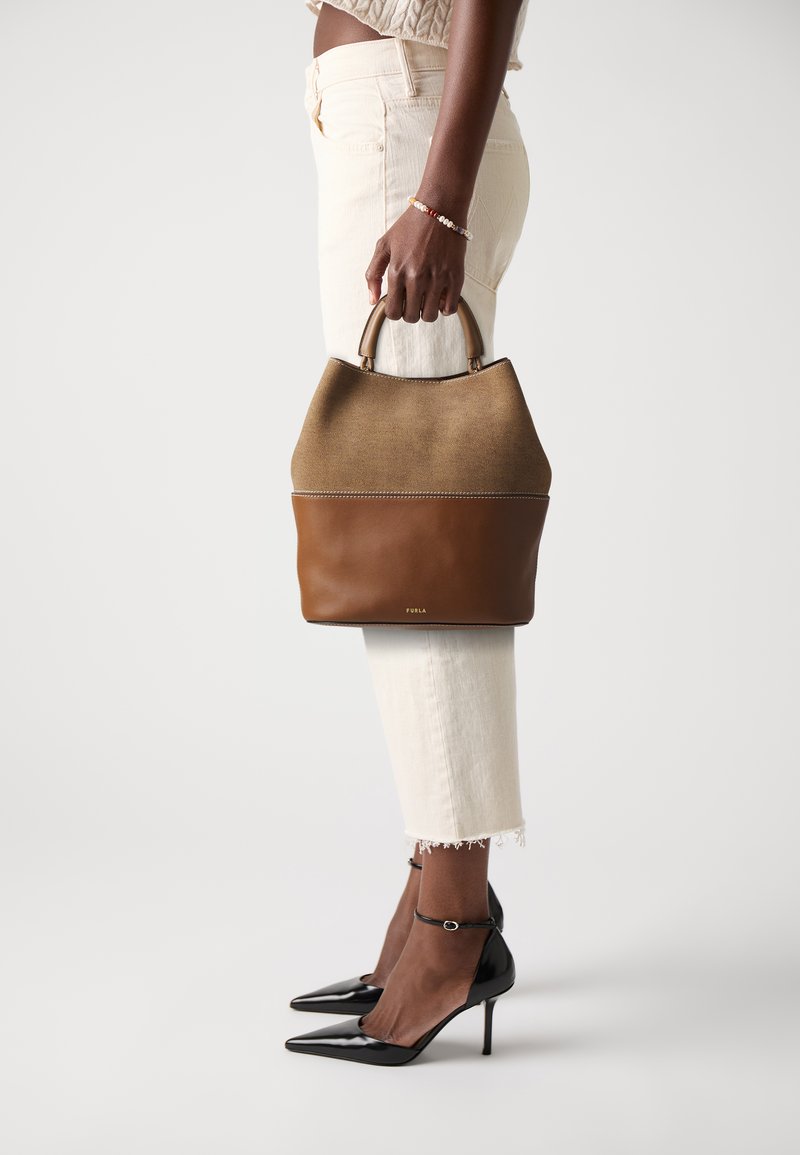 Brown leather handbag with a tan fabric top, curved shape, and contrasting textures; worn with white cropped pants and black high heels.