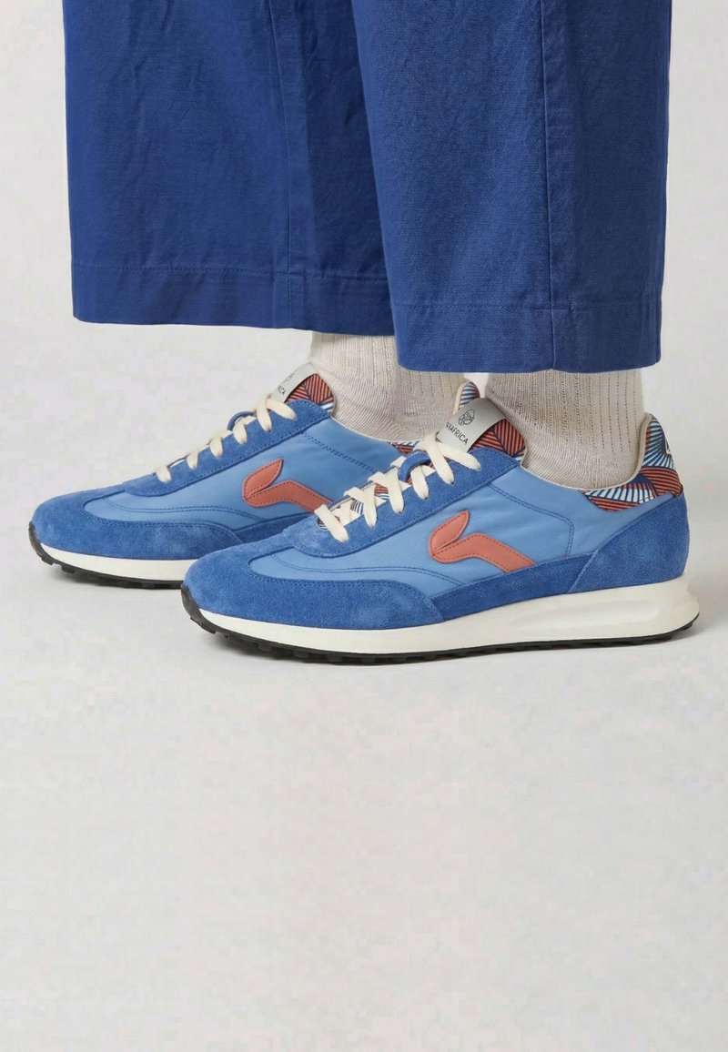 Baskets décontractées bleues et rouges avec des lacets blancs, portées avec des chaussettes beiges et un pantalon bleu raccourci sur un fond clair.