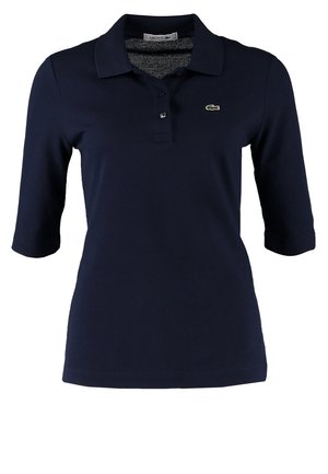 Persona che indossa una polo nera a maniche tre quarti, jeans skinny scuri, sneakers bianche e grigie, portando uno zaino nero su una spalla.
