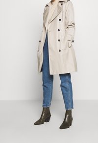 Beżowy trencz o dwurzędowym kroju, z czarnymi guzikami i bocznymi kieszeniami, zestawiony z niebieskimi dżinsami i zielonymi butami na obcasie.