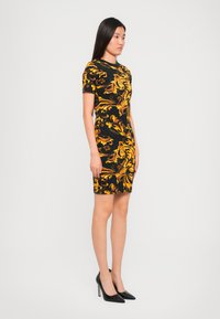 Zwarte jurk met gele barokprint, korte mouwen, getailleerde silhouet, knielange zoom, gecombineerd met zwarte stilettos.