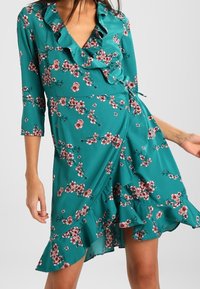 Türaus Kleid in Petrol mit dreiviertel Ärmeln, rüschigem Ausschnitt und floralem Muster in Rosa und Weiß. Mit ausgestelltem Saum und weichem Stoff.