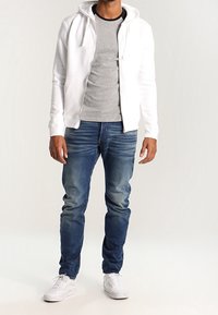 Biała bluza na zamek, noszona na szarej koszulce, w połączeniu z niebieskimi dżinsami i białymi trampkami. Gładki materiał, luźny krój, widoczne detale kaptura.