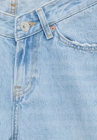 Jean en denim bleu clair avec braguette zippée, fermeture par bouton argenté et surpiqûres jaunes contrastantes. Détail de la poche avant visible.