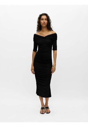 Kvinde med krøllet hår iført en sort off-shoulder midi-kjole med rynker og sorte sandaler mod en ensfarvet lys baggrund.