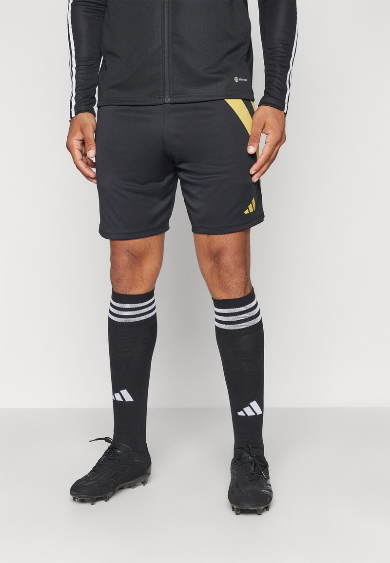 Schwarze Sportshorts mit goldenen Akzenten, kombiniert mit schwarzen knielangen Socken mit weißen Streifen und Logo sowie schwarzen Fußballschuhen.