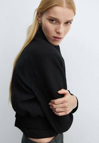 Sort, cropped sweatshirt med høj hals, struktureret stof, elastisk talje og en glat overflade. Modellen poserer med armene korslagt.