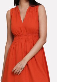 Robe orange sans manches avec un décolleté en V, une taille smockée et un tissu doux. Comprend une jupe fluide et une texture subtile. Bracelet simple en chaîne au poignet.