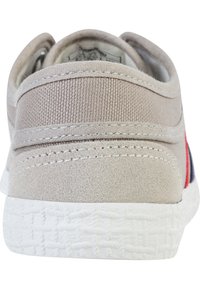 Canvas schoen in lichtbeige, met een textuur achterpaneel, rode en blauwe accenten, en een witte gehudde rubberen zool.