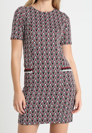 Femme portant une robe à manches courtes avec des motifs géométriques rouges, blancs et noirs et deux détails décoratifs de poche.