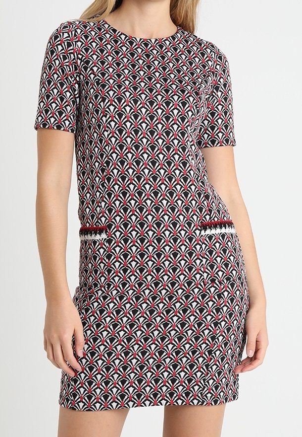 Femme portant une robe à manches courtes avec des motifs géométriques rouges, blancs et noirs et deux détails décoratifs de poche.