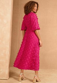 Rochie fucsia cu aplicații florale texturate, mâneci puff scurte și lungime midi, asortată cu sandale cu toc bloc aurii.