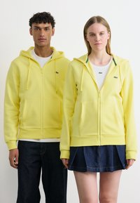 Homme et femme debout côte à côte portant des sweats à capuche assortis jaunes avec fermeture éclair, des hauts blancs, des pantalons foncés et une jupe en denim.