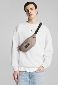 Riñonera color marrón con una superficie texturizada y una etiqueta negra con el logo, llevada de manera diagonal sobre un sudadera blanca, con una correa ajustable negra.