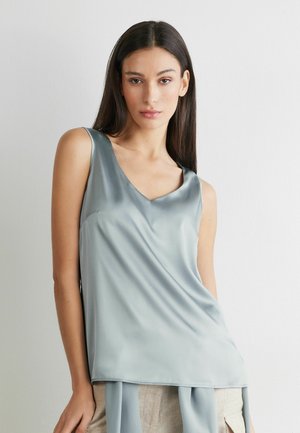 Frau mit langen dunklen Haaren, die ein ärmelloses hellblaues Satintop und beigen Hosen trägt, steht vor einem hellen Hintergrund.