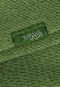 Grüner gestrickter Stoff mit einer strukturierten Oberfläche, versehen mit einem gestickten Etikett in einem dunkleren Grün mit der Aufschrift "spitzbub studios" in weißer Schrift.