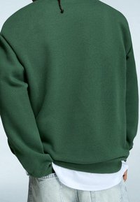Sudadera verde con un corte relajado, puños y dobladillo de canalé. Debajo se ve una camisa blanca. La tela parece suave y texturizada.