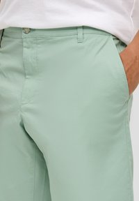 Shorts vert clair en tissu lisse et léger, avec une fermeture à bouton et des poches latérales. Présente un ajustement personnalisé et un devant plat.