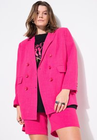 Klart rosa dubbelknäppt blazer med stora knappar, i kombination med matchande shorts. Tyget har en strukturerad yta. Svart grafisk t-shirt under.