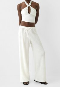 Bílý zkrácený top na ramínka s detaily klíčové díry, v kombinaci s volnými, texturovanými bílými kalhotami s elastickým pasem a stahovací šňůrkou.