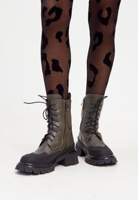 Bottes vert olive à lacets avec une semelle épaisse noire, dotées d'une accent de fermeture éclair et d'un capot de orteil texturé. Portées avec des collants noirs à imprimé léopard.