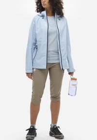 Helleblaue, wasserdichte Jacke mit Kapuze, Reißverschluss und Seitentaschen, kombiniert mit beigen Caprihosen und schwarzen Wanderschuhen, während sie eine Wasserflasche hält.