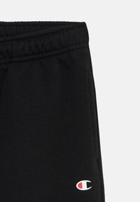 Schwarze Jogginghose aus Baumwolle mit einem elastischen Bund. Mit einem weißen und roten Emblem am unteren linken Bein. Weicher Stoff.