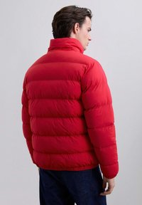 Hombre visto de espaldas vistiendo una chaqueta acolchada roja brillante con cuello alto y jeans azul oscuro, contra un fondo liso.