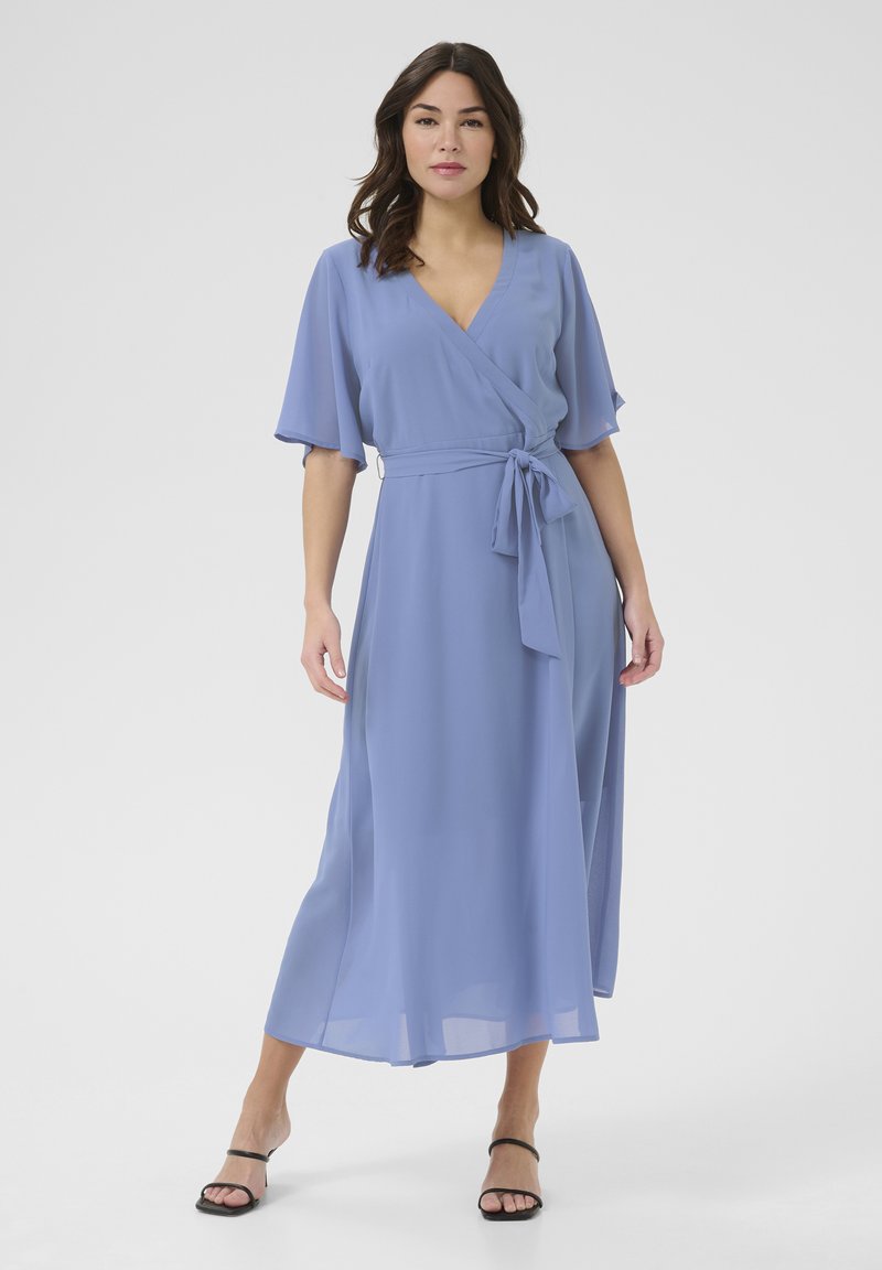 Vestido envolvente azul claro hecho de tela fluida, con mangas cortas acampanadas, un cinturón atado en la cintura y una falda de largo medio. Sandalias negras con tiras.
