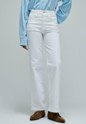 Personne portant un jean blanc taille haute droit, une chemise à manches longues rayée bleu et blanc, et des chaussures en daim marron avec boucles.