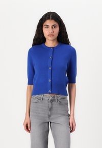 Cárdigan azul corto con mangas cortas, bordes acanalados y cierre de cinco botones. Textura suave, exhibido con jeans de talle alto grises.