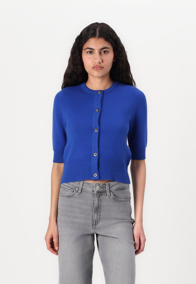 Cárdigan azul corto con mangas cortas, bordes acanalados y cierre de cinco botones. Textura suave, exhibido con jeans de talle alto grises.