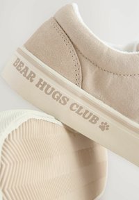 Beige Leinenschuh mit einem strukturierten Obermaterial, weißer Gummisohle und dem auf der Seite aufgedruckten Text "BEAR HUGS CLUB".
