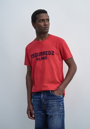 Jeune homme portant un t-shirt rouge DSQUARED2 Milano et un jean bleu, debout avec une main dans la poche, regardant sur le côté devant un fond uni.