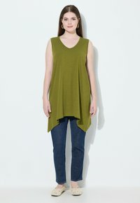 Top senza maniche verde oliva con scollatura a V e orlo asimmetrico, abbinato a jeans blu scuro e scarpe slip-on chiare.