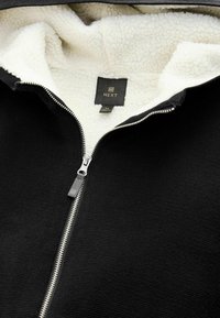 Must mustaga kapuutsiga jakik, millel on tekstureeritud välimus ja pehme valge fliislini, metallne tõmblukk ja brändi silt sees.