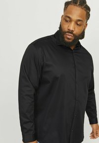 Schwarz longsleeve Hemd mit Button-Down-Kragen, präsentiert mit einer glatten Textur und einer knopfverschlossenen Vorderseite, gezeigt an einer Person vor einem neutralen Hintergrund.