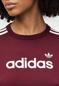 Bordeauxrode katoenen T-shirt met wit "adidas"-logo en klaverbladembleem. Voorzien van een ronde hals en witte gestreepte accenten op de schouders.