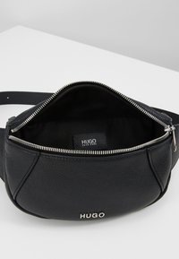 Marsupio in pelle nera con finitura testurizzata, chiusura con zip e interno nero. Presenta un logo argento e dettagli metallici della zip.
