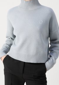 Lichtblauwe coltrui gemaakt van zacht materiaal, met ribbelmanchetten en onderrand, en een klein logo aan de voorkant. Gecombineerd met zwarte broek.