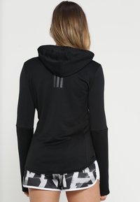 Donna che indossa una maglia nera con cappuccio a maniche lunghe e pantaloni sportivi astratti in bianco e nero, di spalle contro uno sfondo uniforme.