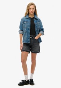 Spijkerjack met twee voorzakken, lichtblauwe kleur, gedragen over een zwart T-shirt. Zwarte denimshorts met rafelige randen, gecombineerd met zwarte schoenen.