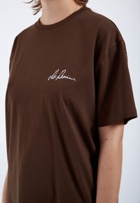 Brown cotton t-shirt with short sleeves, featuring a white embroidered signature on the left side, crew neckline, and smooth texture.