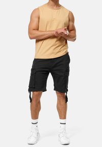 Camiseta sin mangas marrón, pantalones cortos cargo negros con bolsillos laterales y zapatillas blancas con detalles negros. Los pantalones cortos cuentan con correas ajustables en los lados.